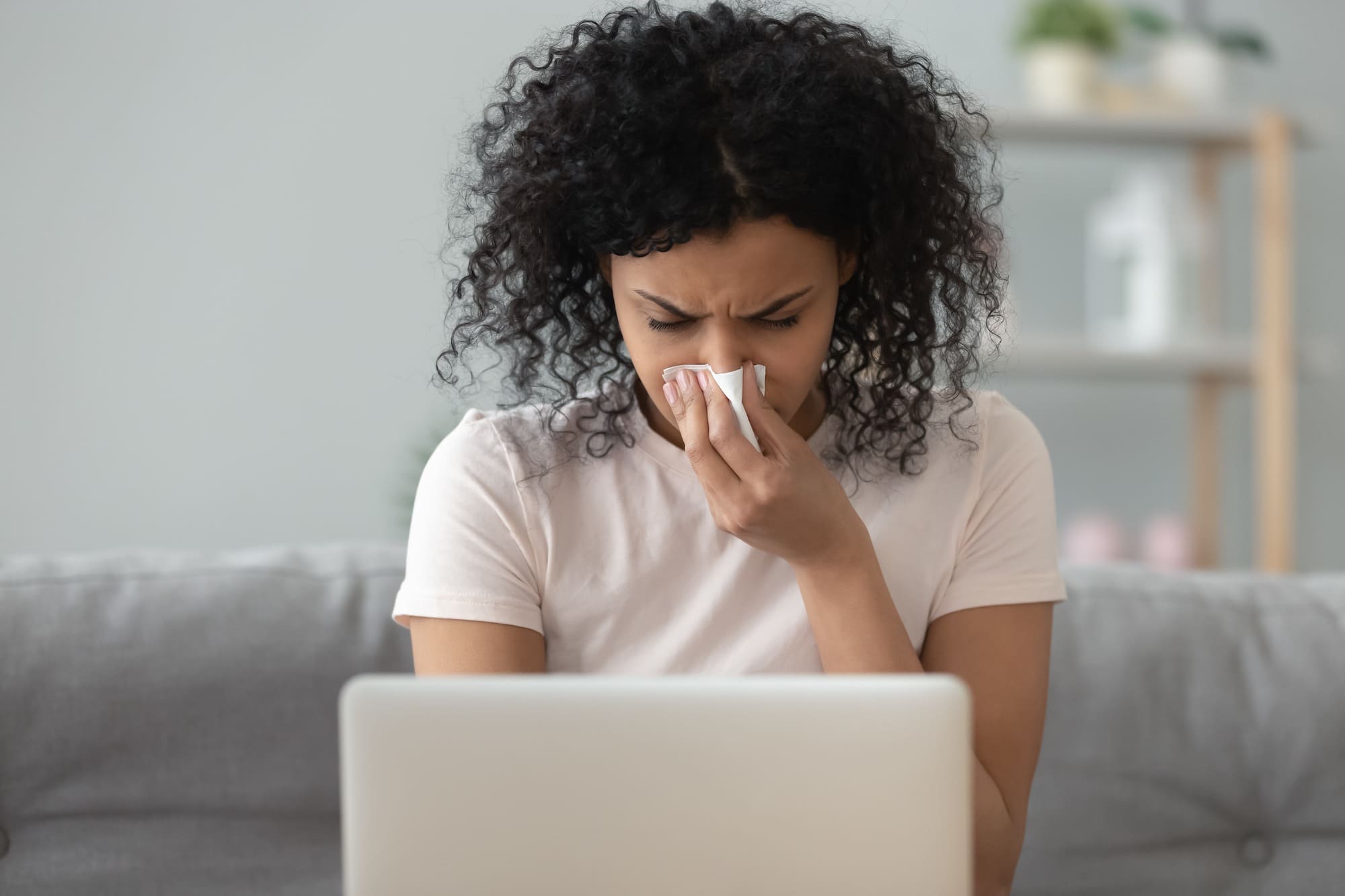 African woman holding handkerchief blowing runny nose working on laptop flu